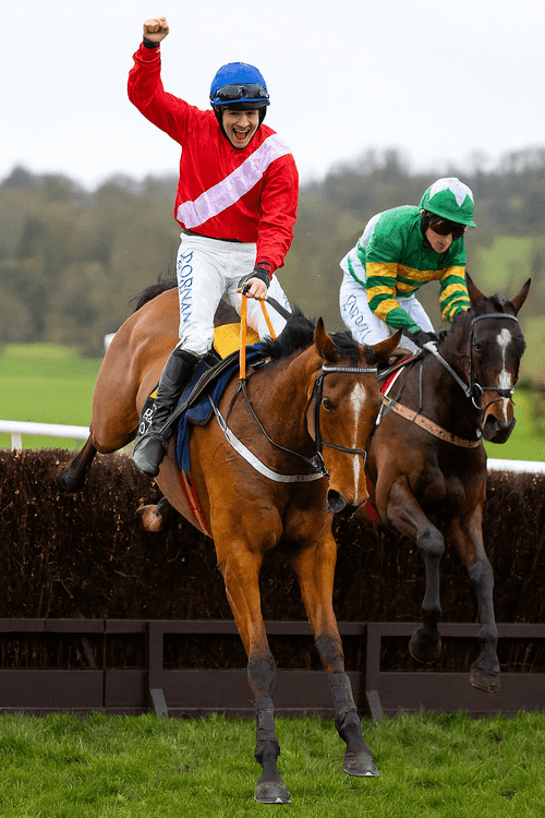 Envoy Allen jumping a fence alongside a rival during a National Hunt chase, ridden by a jockey in red and white silks with a blue helmet, representing Henry de Bromhead’s Knockeen yard.